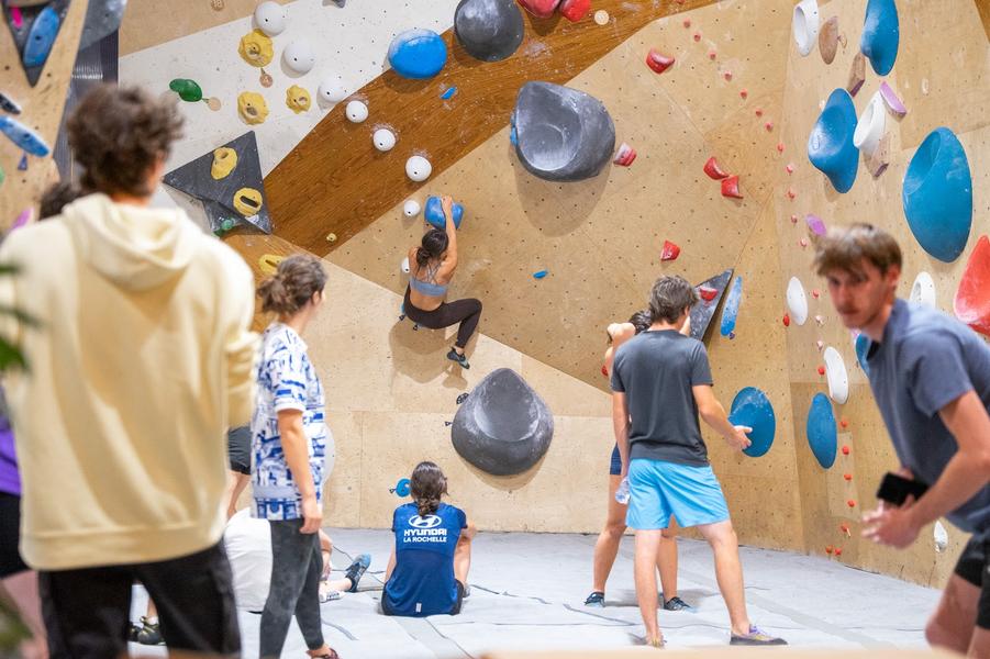 une grimpeuse sur un mur, les autres sur les tapis regardent.